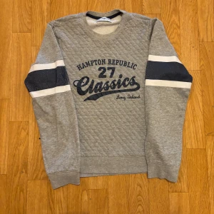 Grå sweatshirt Hampton Republic - Grå quiltad sweatshirt från Hampton Republic med tryck på bröstet och mörkblå samt vita ränder på ärmarna. Klassisk crewneck-modell med lång ärm och mjukt material, perfekt för en avslappnad stil.