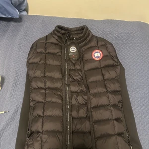 Canada goose väst storlek S, står Storlek M men passar som S. - Säljer en svart dunväst från Canada Goose med quiltad design och klassisk logotyp på bröstet. Västen har hög krage, dragkedja framtill och två sidofickor med dragkedja. Perfekt att slänga över en hoodie för extra värme.