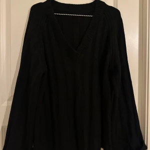 Svart ribbad stickad tröja v-ringad - Svart stickad tröja med v-ringning och ribbade detaljer. Långärmad modell med lite loose fit och breda muddar vid ärmslut. Tillverkad i en mjuk syntetblandning som känns skön mot huden. Perfekt för lager-på-lager under kyliga dagar.