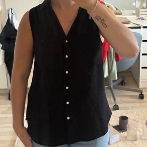 Topp i linne - Svart ärmlös blus med v-ringning och klassisk krage. Toppen har snygga guldiga knappar framtill och två dekorativa bröstfickor. Helt oanvänd 