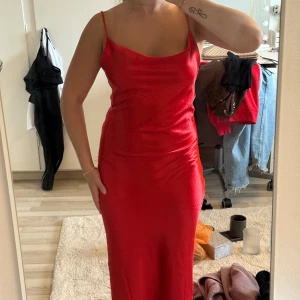 Klänning röd satin - Säljer en röd långklänning i glansigt material med tunna axelband och snygg öppen rygg med dekorativa band. Passar även XS