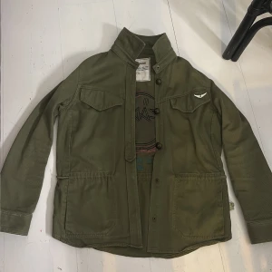 Olivgrön overshirt från Zadig & Voltaire - Snygg olivgrön tunn jacka från Zadig & Voltaire med coolt tryck på ryggen och företagets  vinge på bröstfickan. Jackan har knappar framtill, och den har använts några gånger men inte alls så ofta. Säljer för jag har ingen nytta av den och använder den inte lika mycket. Men en skit cool snygg jacka från Zadig 