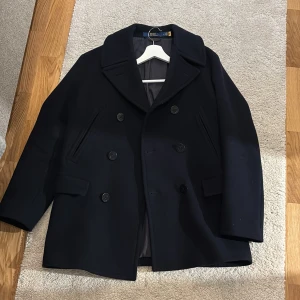 Mörkblå skepparkavaj från Polo Ralph Lauren - Klassisk mörkblå skepparkavaj från Polo Ralph Lauren med dubbelknäppning, bred krage och två fickor fram. Jackan är tillverkad i ull och har en stilren, tidlös look som passar perfekt till höst och vinter. Snygg och enkel att matcha med det mesta.Den är använd en gång, då ny pris på den är sex tusen,priset är inte hugget i sten, det är bara att skicka om det är några funderingar 