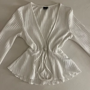 Vit omlottopp med knyt och ribbad struktur - Supersnygg vit omlottopp från Stradivarius med långa ärmar och knyt i midjan. Toppen har ribbad struktur och v-ringning som ger en fräsch och trendig look. Perfekt att styla med jeans eller kjol för en cool vibe.