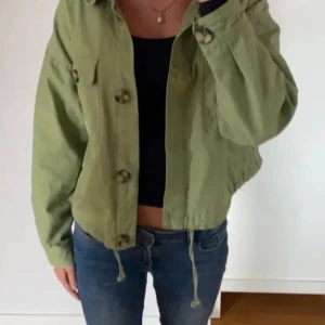 Grön overshirt med stora knappar från bershka  - Snygg ljusgrön overshirt i bomull med stora knappar framtill och två bröstfickor. Jackan har en croppad passform och dragsko nertill för en cool, avslappnad look. Perfekt att slänga över en t-shirt eller hoodie. Passar S-M