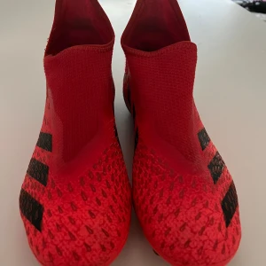 Röda adidas fotbollsskor utan snörning - Snygga röda fotbollsskor från adidas med svarta detaljer och mönstrad ovandel. Skorna har en strumpliknande passform utan snörning och dobbar på sulan för bästa grepp på planen. Perfekta för dig som vill sticka ut på matchen.