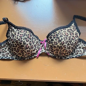 Leopardmönstrad bh med rosa rosett - Jättegullig BH från Triumph som jag säljer eftersom den är för stor för mig. Det finns tyvärr ingen storlek på den, men på den andra bilden visas den på en docka med underbyst 75 cm och byst 88. På tredje bilden har jag stoppat i lite fyllning och då mäts bysten ~93 cm. Så jag skulle gissa den passar någon med underbyst ~70-75 cm och med ett bystmått runt 93cm.