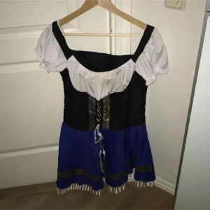 Blå och svart kortklänning med snörning - Säljer en snygg kortklänning i blått, svart och vitt med puffiga kortärmar och snörning framtill. Klänningen har dekorativa gulddetaljer och fransar längst ner på kjolen. Perfekt för temafest eller oktoberfest! Passar dig som vill sticka ut.