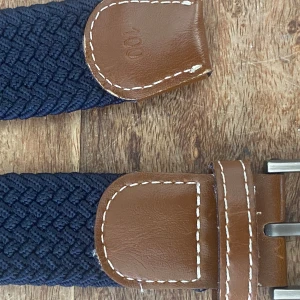 Mörkblå flätat bälte med skinn - Nyköp pris 350kr. Snyggt mörkblått flätat bälte med detaljer i brunt skinn och vita sömmar. Bältet har en klassisk metallspänne och en stilren design som passar till jeans eller chinos. Perfekt accessoar för att lyfta din outfit med en modern touch.