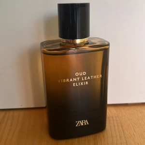 ZARA Oud Vibrant Leather Elixir - En stilren parfymflaska i glas med mörkbrun till svart toning och gulddetaljer. Doften heter Oud Vibrant Leather Elixir och har en lyxig känsla. Flaskan har en svart, blank kork och guldtext på framsidan.