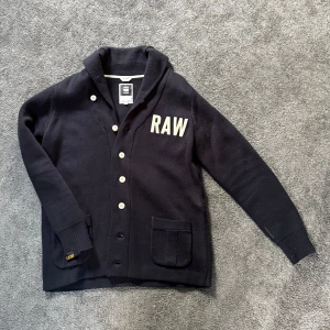 Kofta från G-Star RAW - Sjukt snygg och unik mörkblå kofta från G-Star RAW med stora vita RAW-bokstäver på bröstet. Koftan har en liten sjal krage, vita knappar framtill och två mindre fickor. Står att den är XL men passar precis som L, en person som pendlar mellan M å L passar säkert också i den:) köpte denna för länge sen för drygt 2000kr och den är knappt använd. Ytterst få tecken av användning. Har du någon fråga förutom bild med koftan på då den inte passar så är det bara att fråga på!