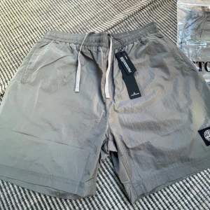 Grå shorts från Stone Island - Snygga grå shorts från Stone Island med klassisk logopatch på benet. Modellen har elastisk midja med snörning och två sidofickor. Materialet är lätt och känns fräscht, perfekt för varma dagar. Kommer i originalförpackning med märkning. Pris kan diskuteras vid snabb affär 