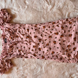 Rosa blommig kortklänning från BikBok - Supersöt rosa kortklänning från BikBok med små orange blommor och knyt vid axeln. Klänningen har en lätt rynkad struktur och är perfekt för varma dagar. Tillverkad i mjukt bomullstyg och har en feminin, somrig vibe.