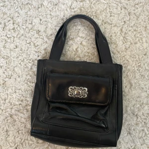 Svart handväska i skinn från Guess - Snygg svart handväska från Guess i äkta skinn med korta handtag. Väskan har en dekorativ silverfärgad detalj på framsidan och ett praktiskt ytterfack med magnetlås. Invändigt finns dragkedjefack för småsaker. Perfekt storlek för det viktigaste.