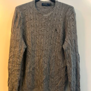 Grå kabelstickad tröja Polo Ralph Lauren - Kabelstickad grå tröja från Polo Ralph Lauren i 100% silke. Klassisk rund halsringning, långärmad och med broderad logga på bröstet. Perfekt för dig som gillar en stilren och tidlös look med lyxig känsla.