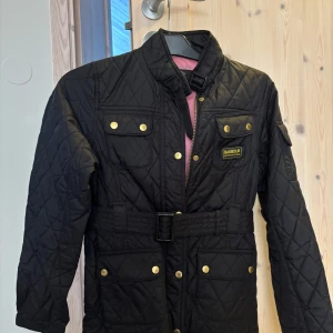 Svart quiltad jacka Barbour XL - Quiltad svart jacka från Barbour International i storlek XL. Jackan har bälte i midjan, guldiga knappar, dragkedja och flera fickor framtill. Klassisk Barbour-patch på bröstet och rosa innerfoder. Perfekt för dig som gillar stilren och sportig look. Inköpt på kidsbrandstore.se därav är storleken XL, mitt namn står inskrivet i jackan. Köparen står för frakten. I mycket fint skick då den bara hängt i garderoben i flera år.