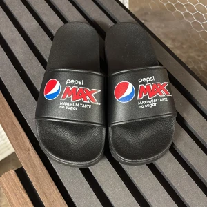 Svarta Pepsi Max sandaler - Svarta sandaler med bred rem över foten och Pepsi Max-logga i rött, blått och vitt. Tillverkade i syntetmaterial med platt sula och öppen tå. Perfekta för chill dagar eller att glida runt i hemma eller på stranden.