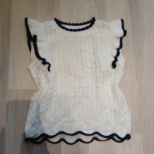 Vit stickad topp med volang från Lindex - Supersöt vit stickad topp från Lindex med mörkblå detaljer. Toppen har volangärmar och vågig kant nertill, samt ett fint hålmönster över hela plagget. Perfekt för dig som gillar en söt och trendig stil! Barnstorlek 170, men passar även storlek 36 eller S i vuxenstorlek. Ej använd, nyskick.