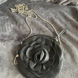 Svart rund axelväska med blomdetalj - Säljer en svart rund axelväska med stor 3D-blomma framtill. Väskan har en silverfärgad kedja som axelrem och dragkedja upptill. Insidan har blommigt mönster och en nyckelring. Liten och smidig väska. Mått 15x15 30kr+frakt