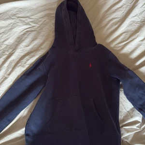 Mörkblå hoodie från Polo Ralph Lauren - Snygg mörkblå hoodie från Polo Ralph Lauren med klassisk röd logga broderad på bröstet. Tröjan har huva, känguruficka och långa ärmar. Perfekt för chill dagar och enkel att matcha med jeans eller joggers. På lappen står det 14-16 år men jag uppskattar den att vara lite mindre från 12-14 år.