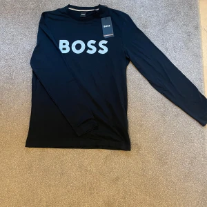 Svart långärmad tröja från BOSS - Helt ny Svart långärmad tröja från BOSS med stor vit logga framtill. Klassisk rund halsringning och stilren design, perfekt för dig som gillar enkel men snygg streetstyle. Tillverkad i mjuk bomull för skön känsla hela dagen.
