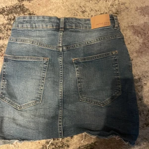 Blå jeanskjol från Perfect Jeans - Snygg blå jeanskjol med rå kant nertill och klassiska fickor både fram och bak. Kjolen har knapp och dragkedja i midjan samt en cool, lite sliten look. Perfekt för dig som gillar en avslappnad och trendig stil.