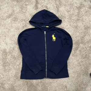 Mörkblå hoodie från Ralph Lauren - Snygg mörkblå hoodie från Ralph Lauren med dragkedja och huva. Tröjan har ett stort gult broderat Polo-logotyp på bröstet och två fickor framtill. Tillverkad i mjuk bomull, perfekt för chill dagar och streetstyle.