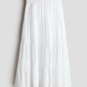 Vit volangkjol med resårmidja - Säljer en vit långkjol med flera volanger och smockad resår i midjan. Kjolen har en luftig och flowy känsla, perfekt för varma dagar. Materialet är mjukt och lätt, troligen bomull. Enkel att matcha med sneakers eller sandaler.