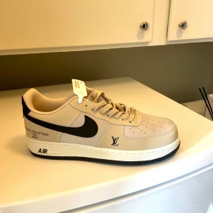 Nike Air Force 1 - Snygga classy beige Nike Air Force 1 sneakers med svarta Louis Vuitton-detaljer på sidan. Skon har klassisk rund tå, snörning och vit sula med svart undersida. Helt nya aldrig använda och kartong och kvitto finns kvar. Storlek US10 som motsvarar storlek 44. Köpta från amonisastore.com för 2569kr.