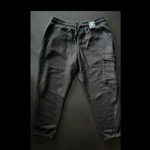 Svarta cargopants från Icono XXL - Svarta cargopants från Icono i storlek XXL. Byxorna har elastisk midja med snörning, stora cargofickor på sidorna och muddar vid bensluten. Tillverkade i mjukt material som är skönt att ha på sig. Perfekta för en chill och avslappnad stil.