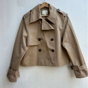 Beige croppad trenchcoat från H&M - Snygg croppad trenchcoat från H&M i beige. Jackan har dubbelknäppning, bred krage, axelklaffar och justerbara spännen vid ärmsluten. Perfekt för dig som vill ha en klassisk men trendig look. Storlek L men passar XS-L beroende på hur oversize man vill ha