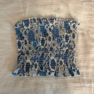 Blommig bandeau tubtopp från Bik Bok - Supersöt bandeau tubtopp från Bik Bok i blått och vitt med blommigt mönster. Smockad och elastisk över hela toppen, med volangkant nertill för extra detalj. Perfekt för varma dagar och enkel att styla med jeans eller kjol.