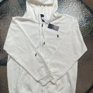 Vit hoodie från Polo Ralph Lauren - Snygg vit hoodie från Polo Ralph Lauren med klassisk broderad logga på bröstet. Tröjan har huva med snörning, dragkedja framtill och ribbade muddar. Tillverkad i mjuk bomull som känns skön mot huden. Perfekt för dig som gillar stilrena och tidlösa plagg. Det är en XL men den är mycket liten i storleken
