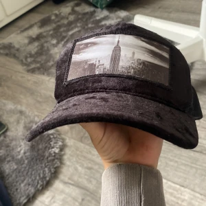 Svart keps med New York-tryck - Snygg svart keps med ett stort tryck av New Yorks skyline och Empire State Building framtill. Kepsen har böjd skärm och justerbar snapback i plast baktill. Materialet är mjukt och har en lätt glansig finish, samt mesh på baksidan för extra ventilation.