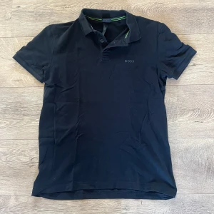 Svart piké från BOSS - Svart piké från BOSS med klassisk krage och korta ärmar. T-shirten har en stilren design med diskret BOSS-logga på bröstet och gröna detaljer på insidan av kragen. Stretchigt material för extra komfort och snygg passform.