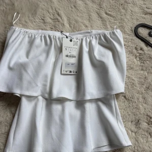 Vit bandeau tubtopp i polyester - Säljer en fräsch vit bandeau tubtopp med volangdetalj upptill. Toppen är axelbandslös och har en mjuk, slät känsla i polyester. Perfekt för dig som vill ha en clean och stilren look till sommaren. Lappen är av dock men super skick💗