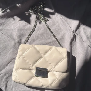 Vit/Beige quiltad axelväska - Snygg vit/beige axelväska från H&M med quiltad design och silvrig kedjerem. Väskan har ett stilrent metallspänne framtill och ett innerfack med dragkedja. Perfekt storlek för det viktigaste och ger en clean look till din outfit.