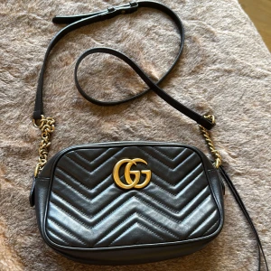 GG Marmont small camera bag - Säljer en svart quiltad axelväska från Gucci med ikoniskt GG-emblem i guld framtill. Väskan är i skinn med kedjedetaljer och justerbar axelrem. Invändigt finns ett ljust foder och praktiska fack. På ena insidans kant finns en bläckfläck. Orginalkvitto från Gucci finns för äkthets garanti.                                                                        Storleken på väskan är: H13, B24, D7 cm 