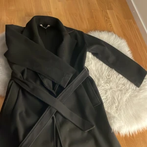 Svart kappa med bälte och krage - Säljer en svart kappa med klassisk krage och bälte i midjan. Jackan har långa ärmar och raka fickor på sidorna. Perfekt för dig som gillar en stilren och clean look. Materialet känns mjukt och har en matt finish.