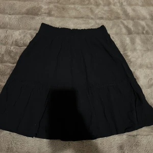 Svart kortkjol - Kjolen har en smockad midja och är tillverkad i ett lätt, skönt tyg med lite struktur. Enkel och stilren modell som är lätt att matcha med olika toppar!