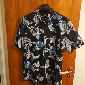 Blommig kortärmad skjorta från H&M - Snygg svart skjorta från H&M med blått och vitt blommigt mönster. Regular fit med korta ärmar och klassisk krage. Perfekt för dig som vill sticka ut med en cool och somrig vibe. Skjortan är i mjuk bomull och har knäppning framtill.