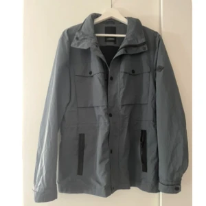 Grå vindjacka från J. Lindeberg - Snygg grön vindjacka från J. Lindeberg med klassisk krage, svarta detaljer vid fickorna och knappar framtill. Jackan har flera praktiska fickor och lång ärm, perfekt för blåsiga dagar och hösten. Enkel och stilren design som funkar till det mesta. Den är i storlek M och är i väldigt fint skick. Kan gå ner i pris vid snabb affär!