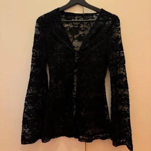 Svart spetsblus med snörning - Supersnygg svart blus i spets med långa utsvängda ärmar och v-ringning. Framsidan har dekorativ snörning som ger en cool och unik look. Perfekt för dig som gillar detaljer och vill sticka ut lite extra.
