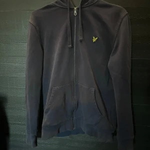 Mörkblå hoodie (Lyle & Scott) - Snygg mörkblå hoodie från Lyle & Scott med dragkedja, huva och snören. Klassisk gul logga på bröstet och ficka framtill.