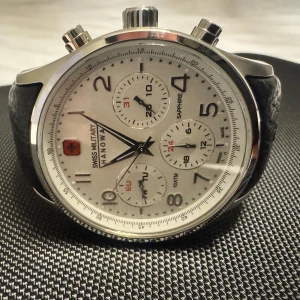 Swiss Military Hanowa kronografklocka - Säljer en helt ny och oanvänd Swiss Military Hanowa chronograph armbandsur. Klockan har en vit urtavla med safirglas och tre sub-urtavlor, en för datumvisning, den andra för veckodagar och den tredje för 24h. Boetten är tillverkad av rostfritt stål och armbandet är tillverkat av äkta läder. Klockan är vattentät till 10 ATM / 100 M. Kommer i originalask.
