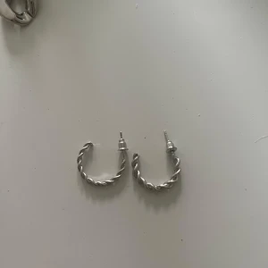 Silvriga tvinnade örhängen - Säljer ett par snygga silvriga örhängen i halvcreol-modell med tvinnad design. De är stilrena och passar perfekt om du vill ha något som sticker ut men ändå är klassiskt. Lätta att matcha med andra smycken.