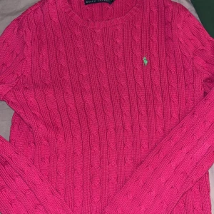 Rosa stickad tröja från Ralph Lauren - Säljer en kabelstickad rosa tröja från Ralph Lauren med rund halsringning och klassisk grön broderad logga på bröstet. Tröjan har lång ärm och är gjord i mjuk bomull, perfekt för lager-på-lager under kyliga dagar.