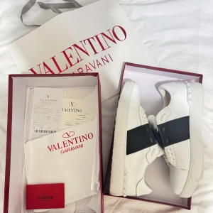Valentino open - Snygga Valentino Garavani Open sneakers i vitt skinn med bred svart rem över vristen och ikoniska nitar på hälen. Klassisk rund tå, platt sula och vita snören. Märkeslogga på plösen i guld. Kommer med originalkartong och dustbag. För mer information, kontakta!