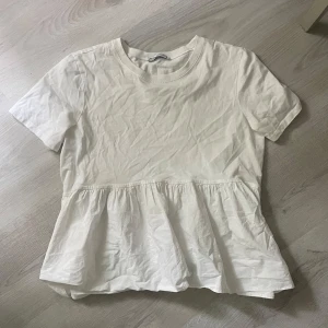 Vit volangblus från Zara - Supersöt vit blus från Zara med volang nedtill som ger en flowy och avslappnad look. Rund halsringning och korta ärmar. Perfekt till jeans eller kjol för en fräsch och enkel stil.