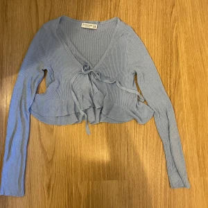 Ljusblå croppad bolerotopp med knyt - Supersöt ljusblå bolerotopp från Stradivarius med ribbad struktur, croppad passform och knyt framtill. Långa ärmar och volangkant nedtill ger en extra gullig vibe. Perfekt att slänga över ett linne eller klänning för en trendig look. Tyget är lite glittrigt.